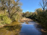A clear stream winding between banks lined with tall trees, a bar of sand and gravel at its edge, blue sky above